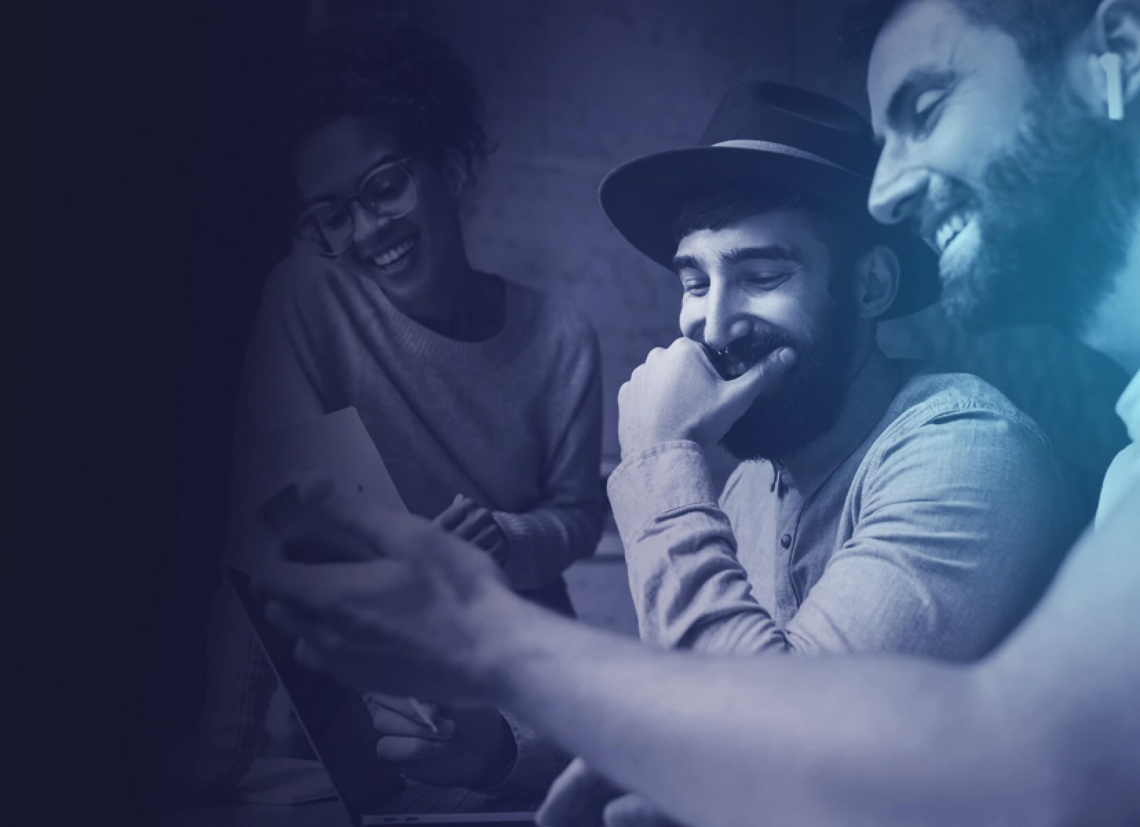 Two adult man sitting, and a woman standing next to them, watching and laughing at something on the phone of one of the men.